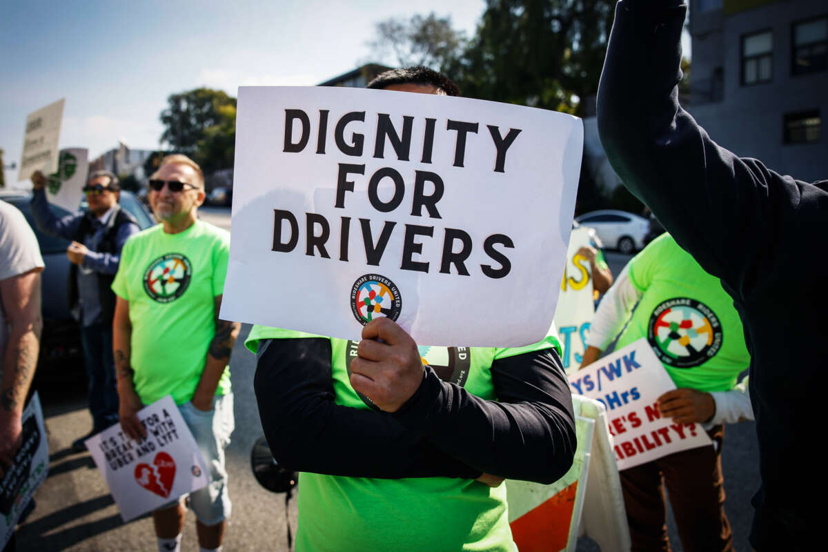 A person in a green shirt holds up a sign reading "DIGNITY FOR DRIVERS" with the Rideshare Drivers United logo beneath the text