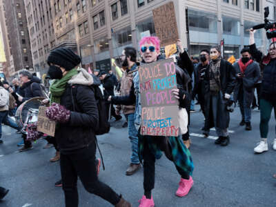 A person with pink hair and matching sneakers holds a sign that reads "POLICE DO NOT PROTECT PEOPLE; THEY PROTECT PROPERTY" during an outdoor protest