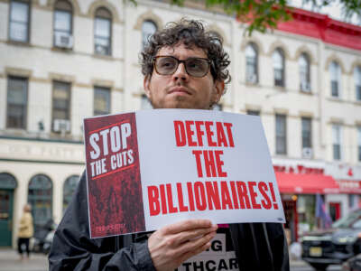 A protester holds a sign reading "STOP THE CUTS; DEFEAT THE BILLIONAIRES" while participating in an outdoor demonstration