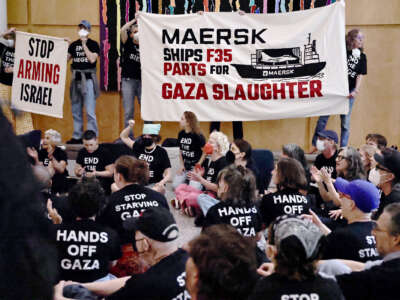 People seated on the ground wearing black shirts with white text that reads "HANDS OFF GAZA", "STOP STARVING GAZA" and "END THE SIEGE" clap and cheer while others hold banners reading "MAERSK SHIPS F35 PARTS FOR GAZA SLAUGHTER" and "STOP ARMING ISRAEL" during an indoor rally