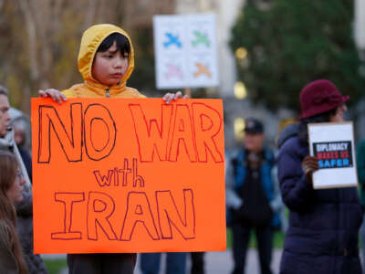 A child in a cute yellow raincoat holds a sign reading "NO WAR WITH IRAN" while in the rain